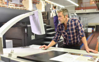 A print shop worker stands at a computer in the workroom, looking intently at the screen.